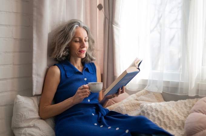 Lovely mature woman in a cobalt blue dress sitting by the window with a cup of tea reading a book - suggestive of self awareness over 40